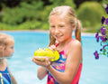 A young girl with blonde hair, wearing a colorful swimsuit, is smiling and playing in a pool. She is holding a green toy water gun shaped like a fish from the Battat Animal Water Squirts collection by Battat, which is squirting water. Another child in a blue swimsuit is visible in the background. Flowers are seen to the right.