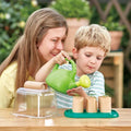 A woman with long hair smiles as she sits beside a young child who is watering seedlings in small pots using a green watering can. Together, they’re engaged in gardening on a wooden table, surrounded by greenery and an eco-friendly *HAPE Gardeners Greenhouse* filled with various gardening supplies from HAPE.