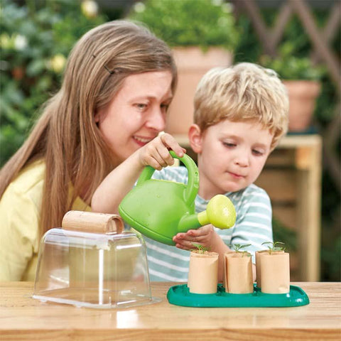 A woman with long hair smiles as she sits beside a young child who is watering seedlings in small pots using a green watering can. Together, they’re engaged in gardening on a wooden table, surrounded by greenery and an eco-friendly *HAPE Gardeners Greenhouse* filled with various gardening supplies from HAPE.