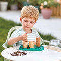 A young child with blond hair, wearing a striped shirt, is sitting at a white table outdoors. They are involved in planting seeds using small pots and a tray filled with soil. The child holds a bamboo paper pot-maker, surrounded by various eco-friendly gardening supplies from HAPE and an *HAPE Gardeners Greenhouse* nearby.