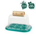 The HAPE Gardeners Greenhouse by HAPE features a plastic container with a green base, six round indentations, and a transparent lid topped with a bamboo handle. It comes with two icons showcasing "Made of Bamboo" and "Made of Plant Plastic," emphasizing its sustainable materials. Ideal for an eco-friendly greenhouse setup.