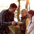 A man, the eco-friendly explorer, demonstrates the HAPE Hand-Powered Flashlight to a young boy and girl in an outdoor setting. Sunlight filters through the trees in the background as the children peer excitedly and curiously at the flashlight's workings.