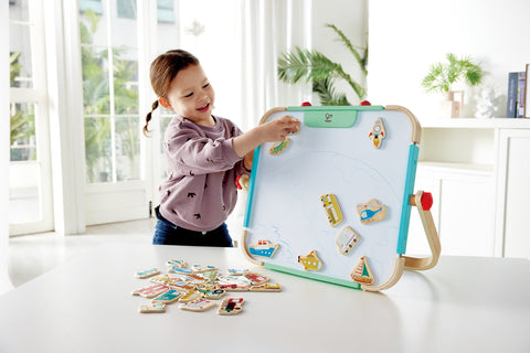 A young girl with a ponytail is playing with her HAPE Magnetic Vehicles 30pc on a table. She is placing the HAPE magnets carefully on the magnetic board, enhancing her fine motor skills. The room is bright and airy, with large windows, plants, and white furniture in the background. Additional HAPE Vehicle magnets are scattered on the table.