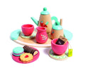 The HAPE Tea Set from HAPE is displayed, featuring a wooden tea service with colorful accents. It includes a teapot adorned with a bird design, a creamer, two teacups, and a sugar bowl, all arranged on a tray. Nearby are two plates with cookies: a green macaron, chocolate roll, and jam-filled biscuit.