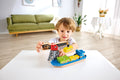 A young child is playing with the Hape Cargo Ship & Crane, which includes a colorful cargo ship and a magnetic crane, on a white table. The background features a green couch, wall art, a plant, and a round white table with a vase of flowers in a bright, modern living room.