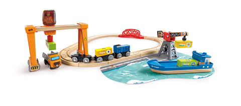 The vibrant Hape Cargo Ship & Crane set by Hape showcases a magnetic crane hoisting a green and yellow container. A blue train with a black engine travels on tracks arranged in an oval, accompanied by a striking red bridge and a gray dock for the blue cargo ship. Additionally, the set includes a small orange truck.