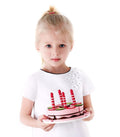 A young girl with blonde hair tied in a ponytail, wearing a white dress with floral accents, holds the Hape Double Flavored Birthday Cake by Hape, which is adorned with three red and white striped candles and colorful decorations. She stands against a plain white background, looking directly at the camera.