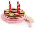 The Hape Double Flavored Birthday Cake by Hape features a toy wooden cake with chocolate icing, decorated with red and white striped candles, green kiwi slices, and red strawberry pieces. The cake is pre-sliced and arranged in a circle on a pink and white plate, complete with felt fruit decorations and a toy wooden serving spatula.