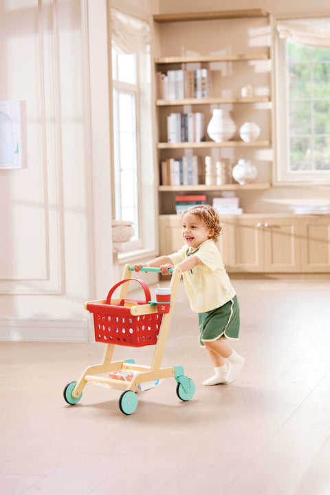 Child playing with a Hape Little Shopper's Cart & Basket in a bright room with wooden furniture.