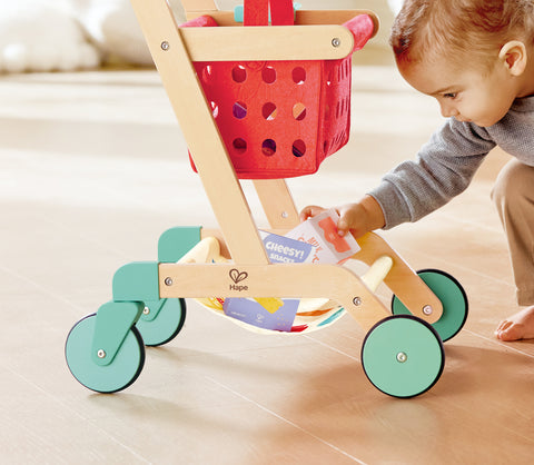 Child playing with a Hape Little Shopper's Cart & Basket on a light wood floor.