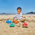 A young child is playing with the Hape Travel Sand Mold Set, featuring a variety of colorful plastic toys and sand molds, on a sandy beach. The set includes a blue boat, an orange watering can, a green sailboat, and a red car. The ocean and distant hills are visible in the background under a clear blue sky.