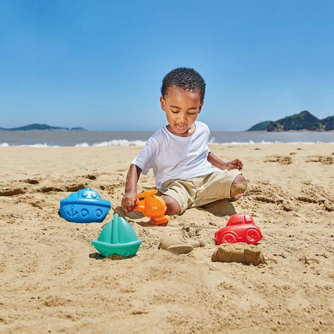 A young child is playing with the Hape Travel Sand Mold Set, featuring a variety of colorful plastic toys and sand molds, on a sandy beach. The set includes a blue boat, an orange watering can, a green sailboat, and a red car. The ocean and distant hills are visible in the background under a clear blue sky.