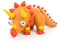 The Hey Clay Single Pack - Triceratops from Hey Clay features a vibrant triceratops model. The dinosaur has an orange body with yellow and red accents, three yellow horns, and large white eyes. Red spikes run along its back and tail, complemented by red feet with yellow patches on its legs, making it perfect for sensory development.