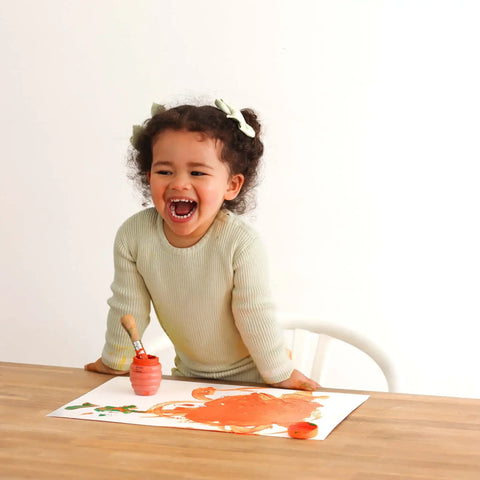 Child laughing while painting on a piece of paper with a brush and red paint.