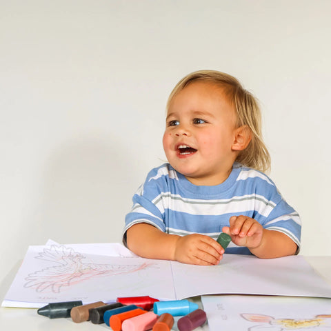 Toddler holding a green Honeysticks crayon and smiling, with a partly coloured page from the Endangered Animals Adventure colouring book on the table