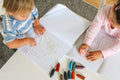 Two toddlers colouring in Honeysticks Toddler's First Colouring Book pages together at a table, with Honeysticks beeswax crayons spread out in front of them