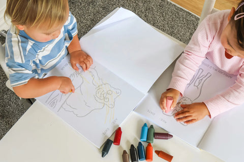 Two toddlers colouring in Honeysticks Toddler's First Colouring Book pages together at a table, with Honeysticks beeswax crayons spread out in front of them
