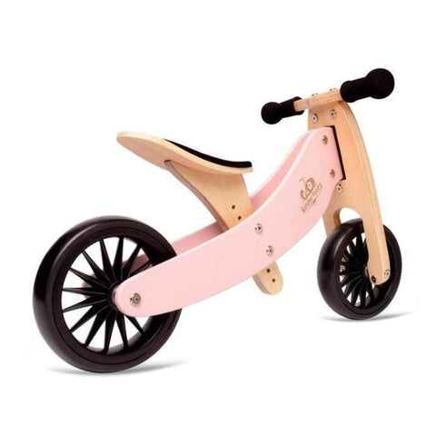 The Kinderboard Tiny Tot Plus - Rose is a children's balance bike featuring a pink wooden frame, black foam handle grips, and black plastic wheels. This 2-in-1 bike boasts a simple, minimalist design with no pedals to help young children learn balance and coordination effortlessly.