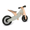 A silver sage Kinderfeet Tiny Tot Plus balance bike by Kinderboard, featuring black rubber tires and designed for young children. The frame combines light wood with gray sections, complemented by black foam grips on the handlebars and a black seat. The intricately designed wheel spokes add a stylish touch.
