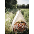 Three young children are sitting inside a small white Kinderboard Kinderfeets Tent in a meadow. Surrounded by tall grass and wildflowers, with a few white daisies near the entrance, they engage in cognitive developmental activities with small baskets and toys, immersed in their fantasy play.