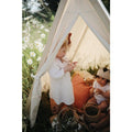 In a sunny meadow adorned with white flowers and greenery, two children are immersed in fantasy play inside a small, white Kinderfeets Tent by Kinderboard. One child, wearing a white dress and a large bow on her head, stands holding something. The other child, sitting in a white outfit and donning a paper crown-hat, engages in activities that foster cognitive development.