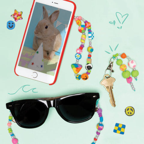 A flat lay image features a smartphone with a rabbit wearing a party hat on the screen, black sunglasses with beaded strings, a key with a colorful handmade beads keychain from Klutz The Ultimate Clay Bead Book by KLUTZ, and scattered stickers including smiley faces, hearts, and peace signs on a light blue background.