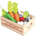 A wooden crate labeled "LE TOY VAN Harvest Vegetables" contains fresh veggies, including green leafy vegetables, carrots with green tops, green beans, a red tomato, white mushrooms, and a halved green vegetable with a darker center. Crafted from sustainable wood, the crate features cut-out handles on the sides.