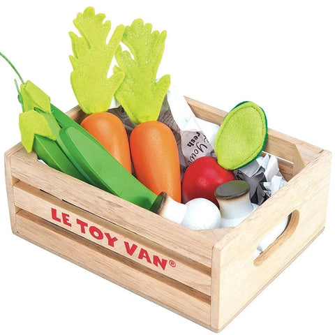 A wooden crate labeled "LE TOY VAN Harvest Vegetables" contains fresh veggies, including green leafy vegetables, carrots with green tops, green beans, a red tomato, white mushrooms, and a halved green vegetable with a darker center. Crafted from sustainable wood, the crate features cut-out handles on the sides.