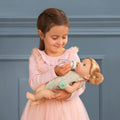 Young girl in a pink dress feeding a Lullababy doll with a bottle against a blue wall.