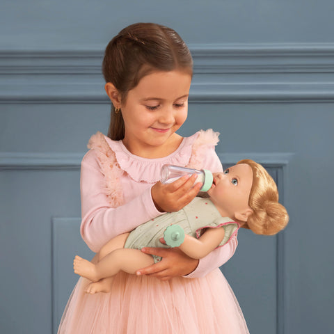 Young girl in a pink dress feeding a Lullababy doll with a bottle against a blue wall.
