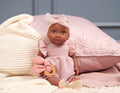 The 14" Lullababy doll is dressed in a pink romper and headband with a bow, sitting against pink quilted pillows and a cream-colored knitted blanket. The dark-skinned doll, part of the Lullababy brand, holds a wooden teething ring with a pink cloth attached. The background features soft, pastel tones.