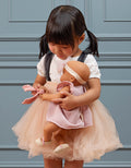 A young girl with dark hair in pigtails stands against a blue paneled wall, wearing a white blouse and a peach-colored tulle skirt. She cradles her Lullababy doll, dressed in the Lullababy 14" Outfit - Pink dress with shoes, gazing down at it affectionately.