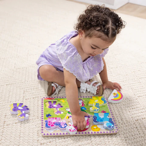 Child sitting on the floor playing with the Melissa & Doug Unicorn Chunky Puzzle, with the rainbow and purple unicorn pieces lifted out beside the board