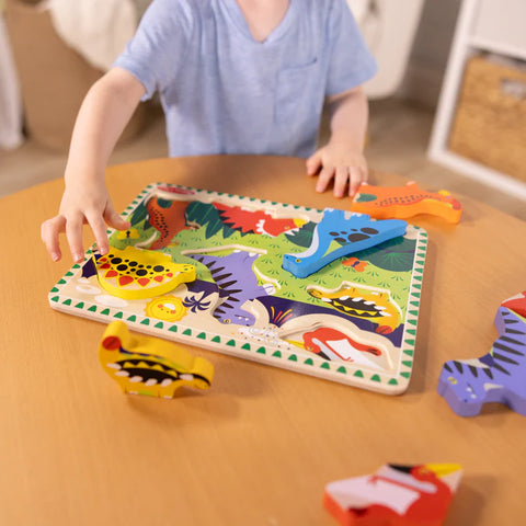 Child reaching for a piece of the Melissa & Doug Chunky Dinosaur Puzzle at a table, with several dinosaur pieces scattered around the board