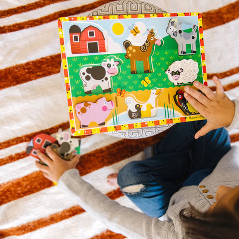 Overhead view of a child sitting with the Melissa & Doug Chunky Farm Puzzle, holding the barn and cow pieces separately beside the board