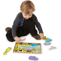 A young child with light brown hair and a grey long-sleeved shirt sits on the floor, deeply engaged in the Melissa & Doug Safari Chunky Puzzle - 8 Pieces. The colorful animal-themed wooden puzzle features chunky wild-animal pieces, including a giraffe, an elephant, a zebra, and a hippo, which help the child develop fine motor skills while they play.