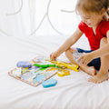 A young child, wearing a red shirt and blue shorts, smiles as they play with the Melissa & Doug Safari Chunky Puzzle – 8 Pieces, featuring colorful wild-animal pieces, on a white bed. The puzzle pieces are scattered around as the child holds one of the pieces, developing their fine motor skills. The background is bright and airy.