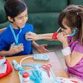 Two children engage in delightful pretend play with the Melissa & Doug Get Well Doctor's Kit Play Set. A girl in a purple shirt uses a toy stethoscope from the set to listen to the chest of a smiling boy wearing a blue shirt. Various toy medical instruments from the Melissa & Doug play set are scattered on the table, with a green sofa as the backdrop.
