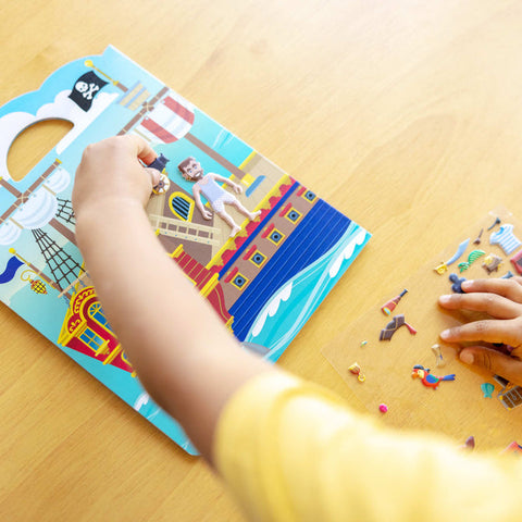 A child wearing a yellow shirt is playing with the Melissa & Doug Puffy Sticker Play Set - Pirate on a wooden table. The child is placing reusable puffy pirate stickers from the Melissa & Doug set onto the board, while additional stickers are spread out nearby, fostering creative expression.