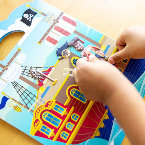 A child is demonstrating their creative expression by assembling the Melissa & Doug Puffy Sticker Play Set - Pirate on a tabletop. The playset features a cartoon pirate ship with a captain figure and vibrant details like sails, flags, and windows. The child's hands are placing a puffy sticker onto the ship's deck.