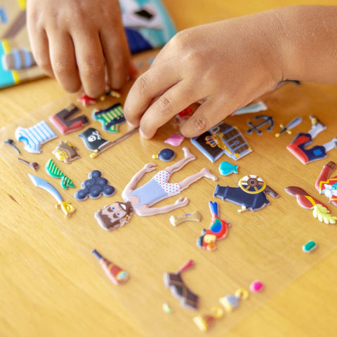 A child’s hands assembling the Melissa & Doug Puffy Sticker Play Set - Pirate on a wooden table with reusable puffy stickers. The stickers include various pirate-themed items such as a pirate figure, cannons, an anchor, a ship’s steering wheel, treasure chests, and other related objects from the Melissa & Doug brand, encouraging creative expression.