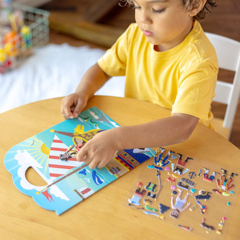 A young child wearing a yellow shirt sits at a round wooden table, engrossed in assembling the Melissa & Doug Puffy Sticker Play Set - Pirate. The play set features pirate characters and a ship, along with reusable puffy stickers for added fun. Several pieces from the set are scattered on the table, while a basket of toys is visible in the background.