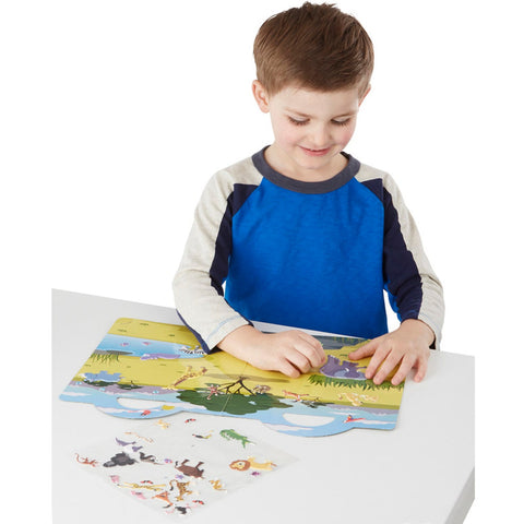 A young boy with light brown hair, clad in a blue and gray long-sleeve shirt, sits at a white table. He is arranging stickers from the Melissa & Doug Puffy Sticker Play Set - Safari on a vibrant play mat that showcases various animal habitats. Next to him, an untouched sheet of puffy animal stickers awaits its turn to join the safari adventure.