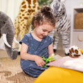 A young child with curly hair sits on the floor surrounded by large stuffed animals, including an elephant, giraffe, and zebra. Wearing a blue dress, the child is engaged with the Melissa & Doug Safari Scissor Skills activity pad from Melissa & Doug, using green scissors to cut paper on a pink surface to develop fine motor skills.
