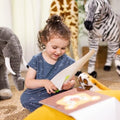 A young girl with curly hair sits on the floor, smiling as she reads a book. Surrounding her are large stuffed animals, including an elephant, giraffe, zebra, and tiger. She wears a patterned blue dress and enjoys her cozy and playful setting while working on Melissa & Doug Safari Scissor Skills projects to enhance her fine motor skills.