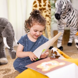 A young girl with curly hair sits on the floor, smiling as she reads a book. Surrounding her are large stuffed animals, including an elephant, giraffe, zebra, and tiger. She wears a patterned blue dress and enjoys her cozy and playful setting while working on Melissa & Doug Safari Scissor Skills projects to enhance her fine motor skills.