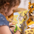 A young child, enhancing their fine motor skills, uses the Melissa & Doug Safari Scissor Skills green scissors to cut colorful paper featuring jungle animals such as lions, tigers, and hippos. In the background, a plush tiger toy can be seen. The child is dressed in a blue shirt with white polka dots.