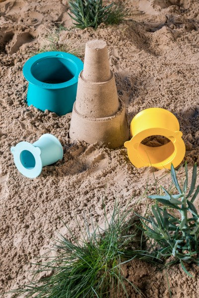A small sandcastle sits on the beach, surrounded by three colorful QUUT Alto molds in blue, yellow, and Lagoon Green. The sand is uneven, with some green plants growing around the molded structures, creating a quaint sand village.