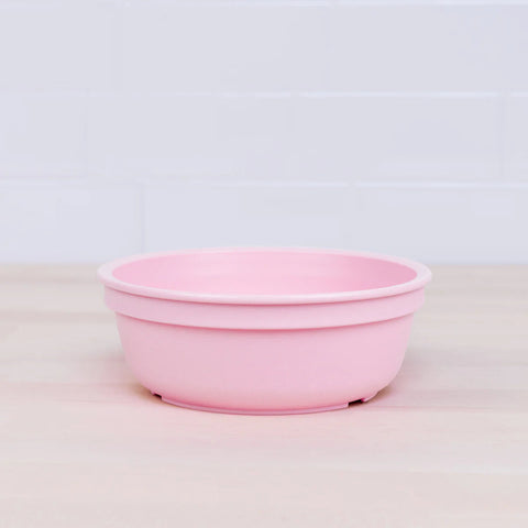 A pink plastic Re-Play Bowl from our vibrant REPLAY tableware set sits empty on a light wooden countertop with a white tile backsplash. The bowl has a simple, smooth design, and its muted pastel color contrasts subtly with the light background, making it both safe for kids and environmentally friendly.