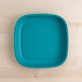 A Re-Play Flat Plate by REPLAY, featuring a square shape with slightly rounded corners in teal, crafted from recycled materials, is placed on a light wooden surface.